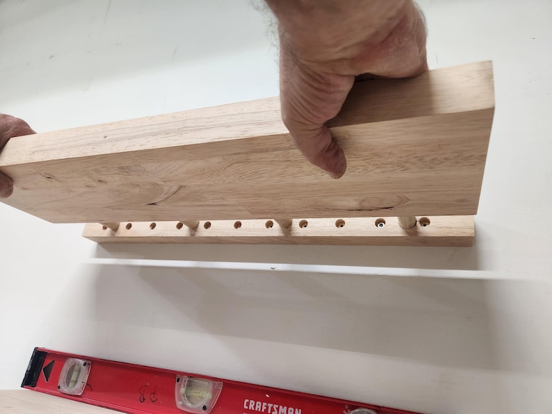 Puede incluir: Un estante de pared de madera de color claro que presenta una fila de orificios circulares espaciados uniformemente a lo largo del borde inferior, con un nivel de burbuja rojo marca Craftsman colocado debajo para asegurar la alineaci&oacute;n.