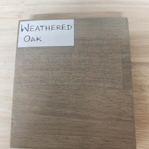 Puede incluir: Una muestra de madera cuadrada con acabado de roble desgastado, que presenta un patr&oacute;n de veta natural visible en tonos grises y marrones. Una etiqueta rectangular blanca est&aacute; pegada en la esquina superior izquierda con el texto Weathered Oak.
