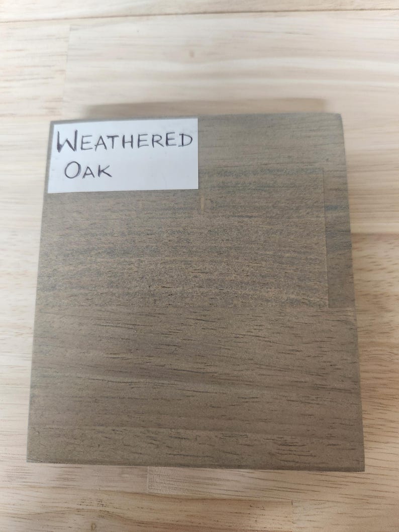 Puede incluir: Una muestra de madera cuadrada con acabado de roble desgastado, que presenta un patr&oacute;n de veta natural visible en tonos grises y marrones. Una etiqueta rectangular blanca est&aacute; pegada en la esquina superior izquierda con el texto Weathered Oak.