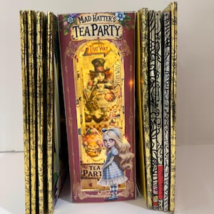 Puede incluir: Una pila de libros con cubiertas de patrones dorados. El libro del centro es un libro desplegable con una cubierta roja y el título "Mad Hatter's Tea Party". El desplegable presenta una escena de Alicia en el País de las Maravillas con Alicia y el Sombrerero Loco.