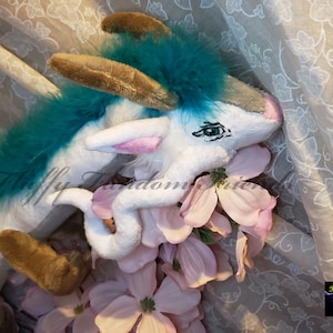 Puede incluir: Un dragón de peluche blanco con plumas azul turquesa, cuernos marrones y orejas rosas. El dragón está enroscado con flores rosas.