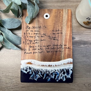 May include: Wooden cutting board with a handwritten recipe for "My Chocolate Pie." The board features a resin wave design in white and blue at the bottom. A mug with a guitar and the word "Dream" is in the background.
