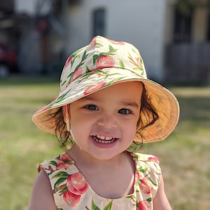 May include: A young child wearing a yellow and green sun hat with a peach print. The hat has a wide brim and a chin strap. The child is smiling and looking at the camera.