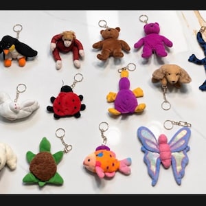 May include: A collection of thirteen plush animal keychains featuring a penguin, monkey, brown bear, purple bear, blue lizard, white bird, ladybug, yellow and purple creature, dog, sheep, green turtle, orange fish, and a blue and purple butterfly.