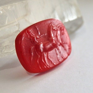 May include: A red carnelian gemstone carved with a detailed image of a horse. The gemstone is oval shaped and has a smooth, polished surface.