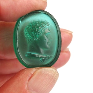May include: Oval, emerald-green glass cameo featuring a profile of a person with curly hair and a draped garment. The cameo is held between fingers against a white background. The glass has a translucent quality.