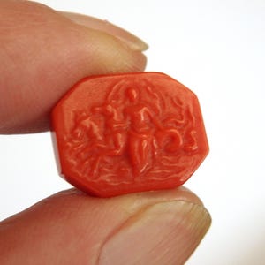 May include: A small, octagonal, coral-red gemstone with a carved relief depicting figures. The carving shows a central figure flanked by others, possibly mythological. The gemstone is held between two fingers against a white background.