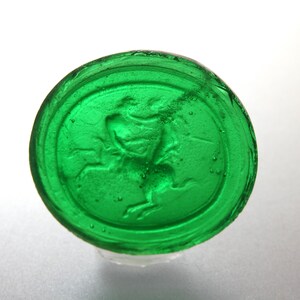 May include: A round, transparent, emerald green glass object with a raised image of a person on a horse. The glass has a slightly textured surface and is set against a white background.