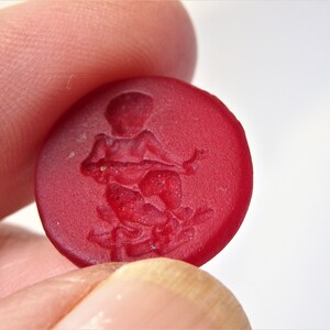 May include: A red glass button with a raised design of a person holding a staff. The design is in a darker shade of red.