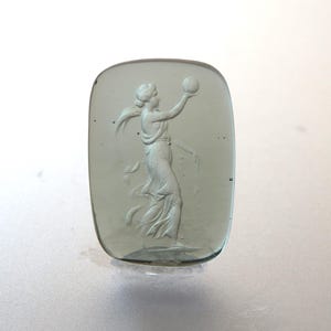 May include: A rectangular, translucent, pale green glass cameo. The cameo features a classical female figure holding a sphere aloft. The figure is draped in flowing robes, with her hair pulled back. The edges of the cameo are rounded.