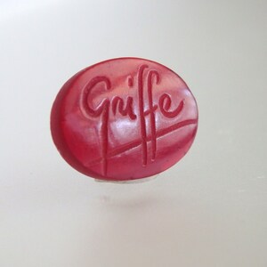 May include: A red oval-shaped button with the word "Griffe" embossed in white.