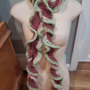 May include: A crocheted scarf featuring a ruffled design with a color palette of brown, light green, and soft pink. The yarn is worked in a wavy, layered pattern that creates a voluminous texture throughout the length of the accessory.