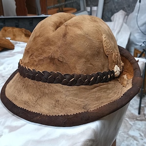 Könnte beinhalten: Brauner Lederhut mit breiter Krempe und geflochtenem Band. Der Hut hat eine strukturierte Oberfläche und dekorative Elemente an der Seite. Der Rand ist mit dunklerem braunem Leder verziert.