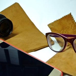 Può includere: Primo piano di un obiettivo di una fotocamera nera, uno smartphone rosso, un paio di occhiali con montatura viola e due panni in pelle scamosciata marroni.