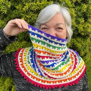 May include: A rainbow-coloured crocheted cowl. The cowl displays a vibrant spectrum of colours, including red, orange, yellow, green, blue, and purple, with a white base. The edges are trimmed with a red scalloped border. The cowl is worn around the neck.