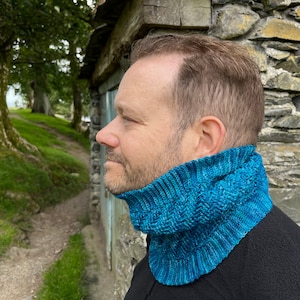 May include: A vibrant teal blue knitted cowl neck warmer featuring a textured ribbed pattern. The accessory is crafted with a variegated yarn that displays subtle shifts in blue and green tones, providing a snug fit around the neck.
