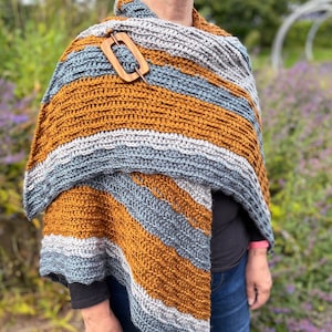 May include: A handmade crochet shawl in shades of gray, mustard yellow, and white, secured with a wooden shawl pin. The striped pattern creates a cozy and stylish accessory, perfect for cooler weather.
