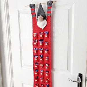 Peut inclure: Un calendrier de l'avent en crochet rouge en forme de gnome suspendu à une porte blanche. Le gnome a un chapeau et une barbe gris, et un cœur blanc pour la poitrine. Le calendrier a 24 poches numérotées avec des boutons en bois.