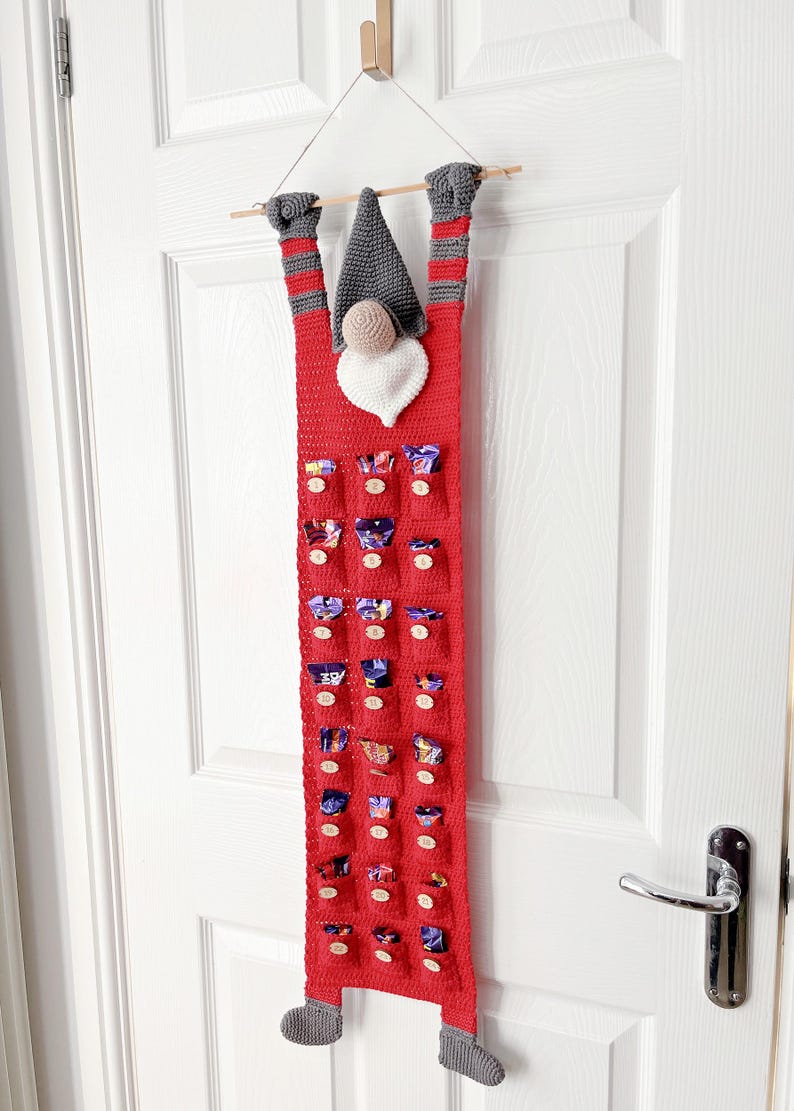 May include: A crocheted advent calendar shaped like a gnome hanging on a door. The gnome is gray and white, with a red body and gray boots. The calendar has 24 numbered pockets filled with small wrapped chocolates. The calendar is made of red yarn and features a charming gnome design. It's a handmade Christmas advent calendar perfect for counting down the days to Christmas.