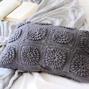 May include: A rectangular, dark gray crochet pillow with a floral pattern. The pillow is made of soft yarn and is placed on a white surface with a gray blanket and an open book in the background. The pillow is approximately 12 inches wide and 20 inches long.