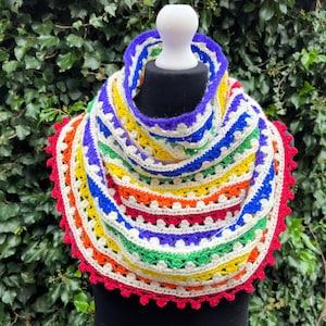 May include: A colorful crocheted cowl with a rainbow pattern and a white crocheted border. The cowl is displayed on a mannequin against a green leafy background.