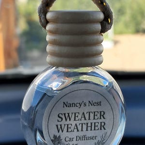 May include: A clear glass car diffuser bottle with a light wood cap and a thick hanging cord. The circular label features the text Nancy's Nest, SWEATER WEATHER, and Car Diffuser, accompanied by a small illustration of pumpkins and pinecones.