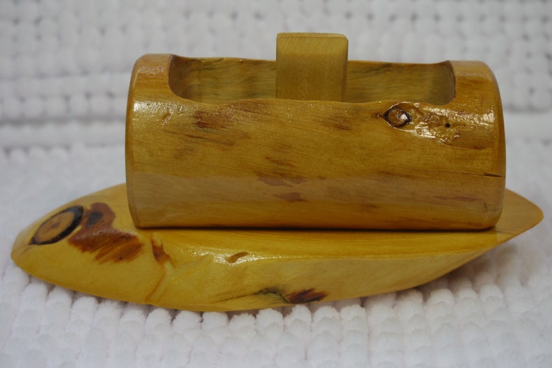 May include: A wooden boat toy with a natural wood finish. The boat has a rounded hull and a small, rectangular sail. The boat is sitting on a white surface.
