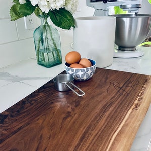 May include: A large, rectangular, wooden cutting board with a live edge. The board is made of dark walnut wood and has a natural, rustic finish. The board is engraved with the text "The Rothman Family".