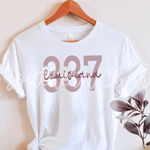 May include: White t-shirt with a pink graphic design featuring the number 337 and the word "Louisiana" underneath.