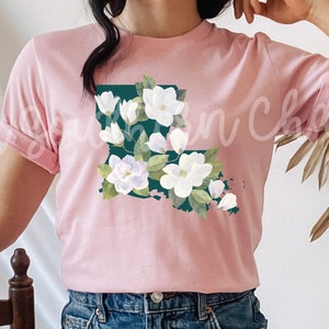 K&ouml;nnte beinhalten: Ein T-Shirt in zartem Rosa mit einem gr&uuml;nen Umriss des Bundesstaates Louisiana, verziert mit wei&szlig;en Magnolienbl&uuml;ten. Der Text "Soft Pink" ist in Schwarz am unteren Rand des Bildes geschrieben.