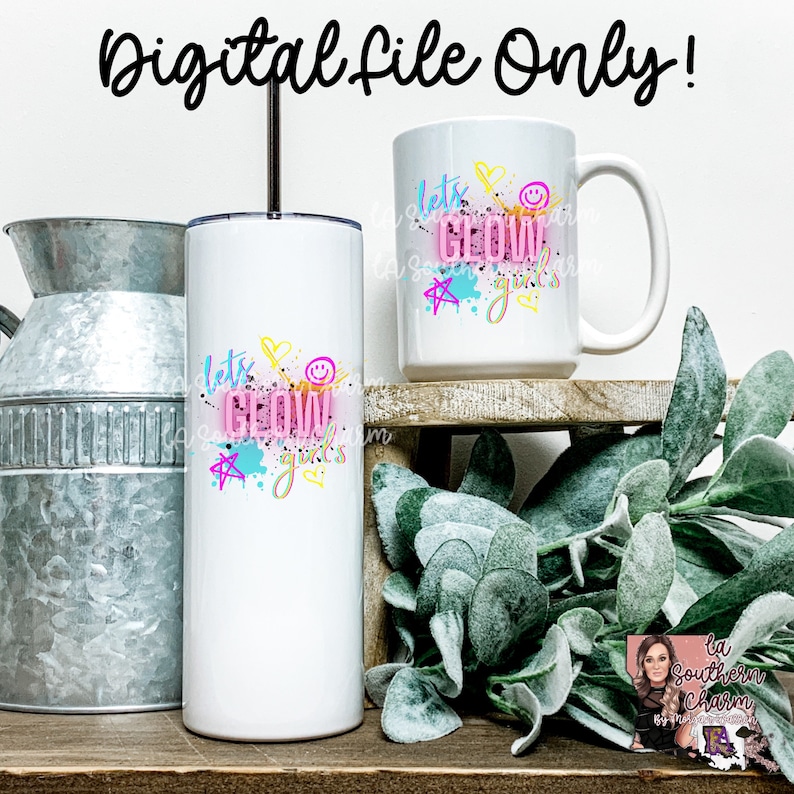 May include: Two white tumblers with a pink, blue, and yellow design that says "Let's Glow Girls". The tumblers are on a wooden shelf with a green plant in the background.