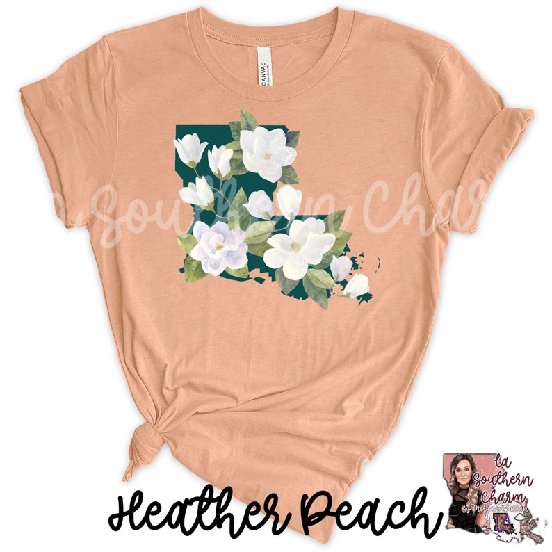 K&ouml;nnte beinhalten: Ein pfirsichfarbenes T-Shirt mit einer Grafik des Bundesstaates Louisiana, das in blaugr&uuml;ner Farbe umrandet und mit wei&szlig;en Magnolienbl&uuml;ten gef&uuml;llt ist. Der Text "Heather Peach" ist auf der Unterseite des T-Shirts aufgedruckt.