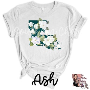 K&ouml;nnte beinhalten: Wei&szlig;es T-Shirt mit einem t&uuml;rkisfarbenen Umriss des Bundesstaates Louisiana, verziert mit wei&szlig;en Magnolienbl&uuml;ten. Der Text "Ash" ist in schwarzer Kursivschrift am unteren Rand des Shirts geschrieben.