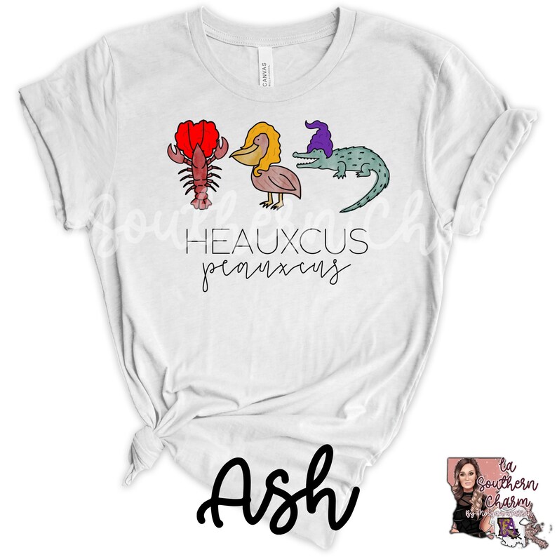 May include: White t-shirt with a cartoon illustration of a lobster, pelican, and alligator wearing a purple hat. The text "Heauxcus Peauxxus" is printed below the illustration.