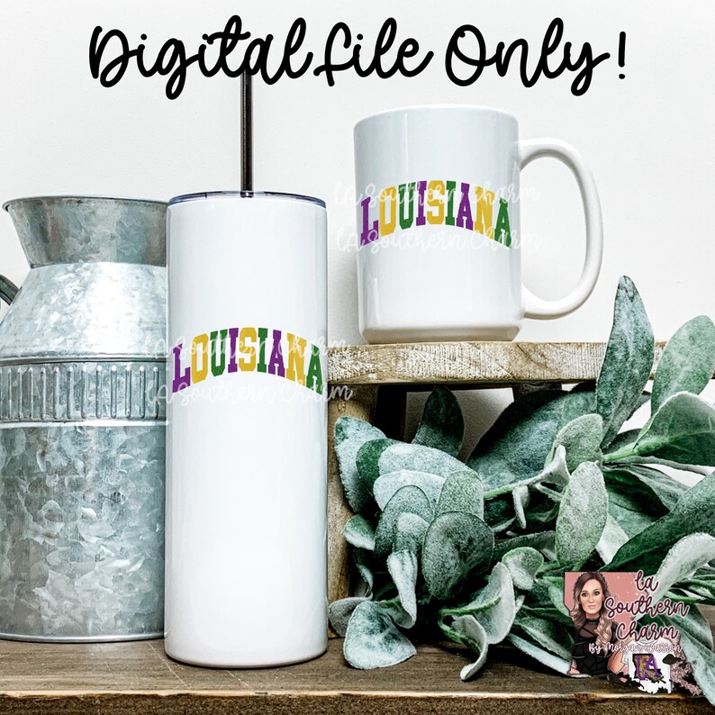 May include: A white tumbler and a white mug with the word "LOUISIANA" in green, purple, and gold letters. The tumbler and mug are on a wooden shelf with green plants.