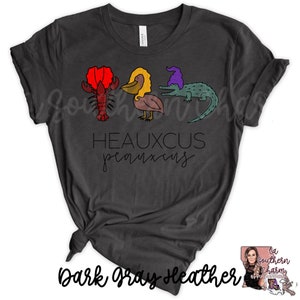 May include: Dark gray heather t-shirt with a graphic of a crawfish, pelican, and alligator wearing a purple hat. The text "Heauxcus  peauxcus" is printed below the graphic.