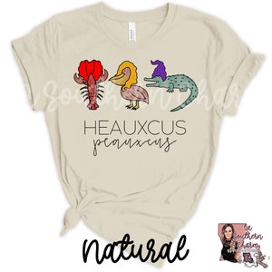 May include: A beige t-shirt with a graphic of a lobster, pelican, and alligator wearing a purple hat. The text "Heauxcus pearlxus" is printed below the graphic. The word "natural" is written in black cursive at the bottom of the shirt.