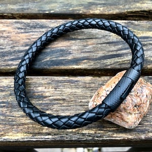 May include: A black leather bracelet with a braided design and a metal clasp. The bracelet is lying on a wooden surface.