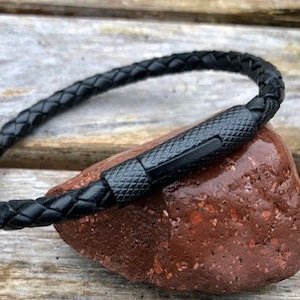 May include: A black braided leather bracelet with a black clasp, resting on a reddish-brown stone. The bracelet is circular and appears to be made of woven leather strands. The clasp has a textured pattern.
