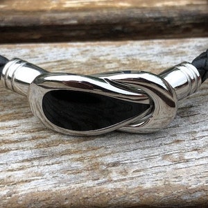 May include: A black leather bracelet with a silver clasp featuring a knot design. The clasp has a black inlay.