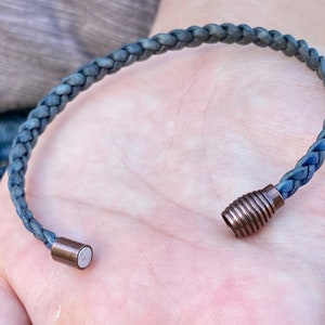 K&ouml;nnte beinhalten: Geflochtenes blaues Armband mit kupferfarbenem Magnetverschluss. Das Armband besteht aus einem gewebten Material und der Verschluss hat eine zylindrische Form. Das Armband wird auf der Hand einer Person gezeigt.