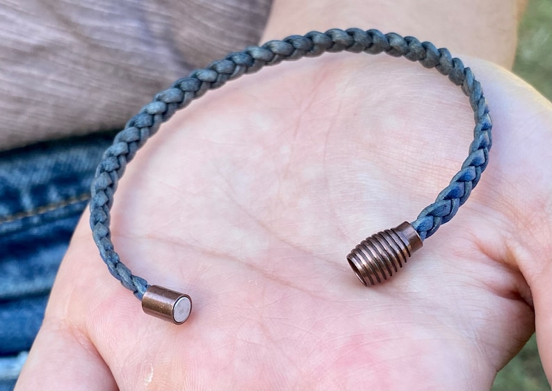 K&ouml;nnte beinhalten: Geflochtenes blaues Armband mit kupferfarbenem Magnetverschluss. Das Armband besteht aus einem gewebten Material und der Verschluss hat eine zylindrische Form. Das Armband wird auf der Hand einer Person gezeigt.