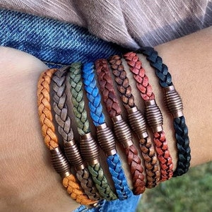 K&ouml;nnte beinhalten: Eine Sammlung geflochtener Lederarmb&auml;nder in verschiedenen Farben, darunter Orange, Grau, Gr&uuml;n, Blau, Braun, Rot und Schwarz. Jedes Armband hat einen kupferfarbenen Magnetverschluss. Die Armb&auml;nder werden an einem Handgelenk gezeigt.