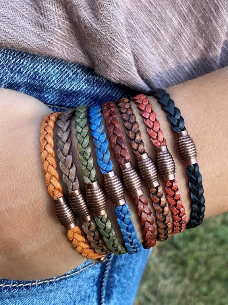 K&ouml;nnte beinhalten: Eine Sammlung geflochtener Lederarmb&auml;nder in verschiedenen Farben, darunter Orange, Grau, Gr&uuml;n, Blau, Braun, Rot und Schwarz. Jedes Armband hat einen kupferfarbenen Magnetverschluss. Die Armb&auml;nder werden an einem Handgelenk gezeigt.