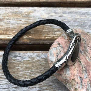 May include: A black leather bracelet with a silver clasp. The clasp is a unique design with a smooth, rounded shape.