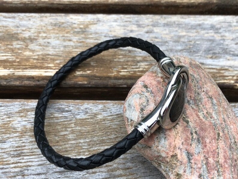 May include: A black leather bracelet with a silver clasp. The clasp is a unique design with a smooth, rounded shape.