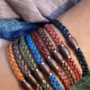 K&ouml;nnte beinhalten: Eine Auswahl an geflochtenen Lederarmb&auml;ndern in verschiedenen Farben, darunter Orange, Grau, Gr&uuml;n, Blau, Braun, Rot und Schwarz. Jedes Armband hat einen Metallverschluss. Modisches Accessoire.