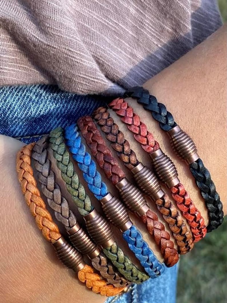 K&ouml;nnte beinhalten: Eine Auswahl an geflochtenen Lederarmb&auml;ndern in verschiedenen Farben, darunter Orange, Grau, Gr&uuml;n, Blau, Braun, Rot und Schwarz. Jedes Armband hat einen Metallverschluss. Modisches Accessoire.