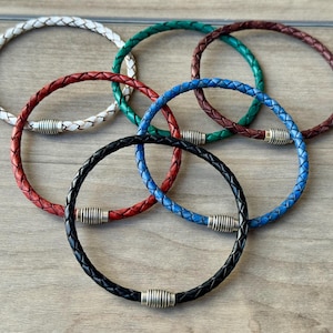 May include: A collection of braided leather bracelets in various colors, including white, red, green, blue, brown, and black. Each bracelet features a silver-toned magnetic clasp. The bracelets are arranged on a wooden surface.