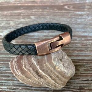 Peut inclure: Un bracelet en cuir tress&eacute; noir avec une fermeture en cuivre. Le bracelet est sur un rocher.